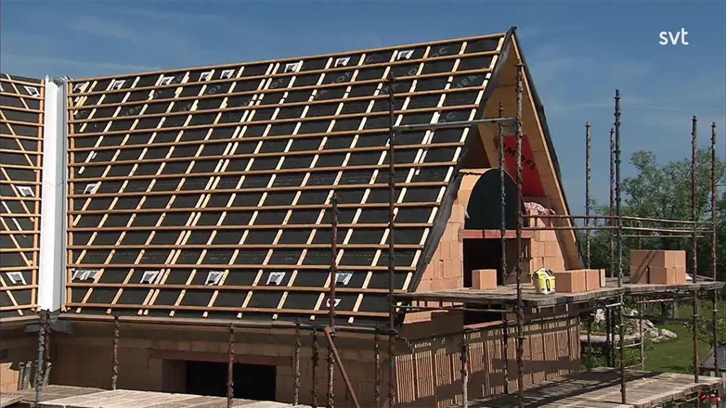 Stiligt - Ett hus med brant lutande tak är under uppbyggnad, med byggnadsställningar runt omkring och träribbor installerade för takläggning.