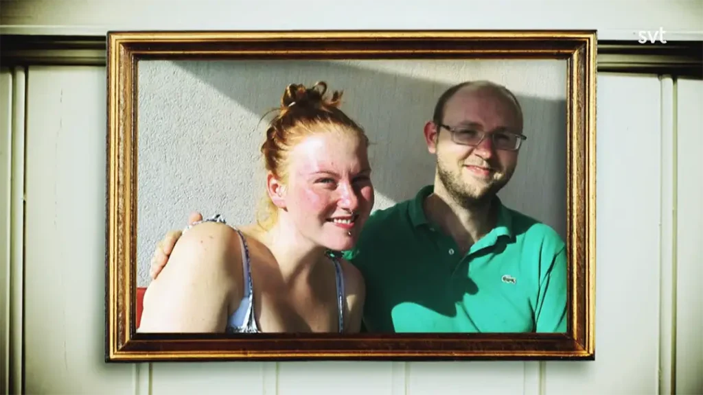 Stiligt - Ett inramat foto av en leende kvinna med rött hår och en man i glasögon och grön skjorta, sittande tätt tillsammans mot en ljus vägg.