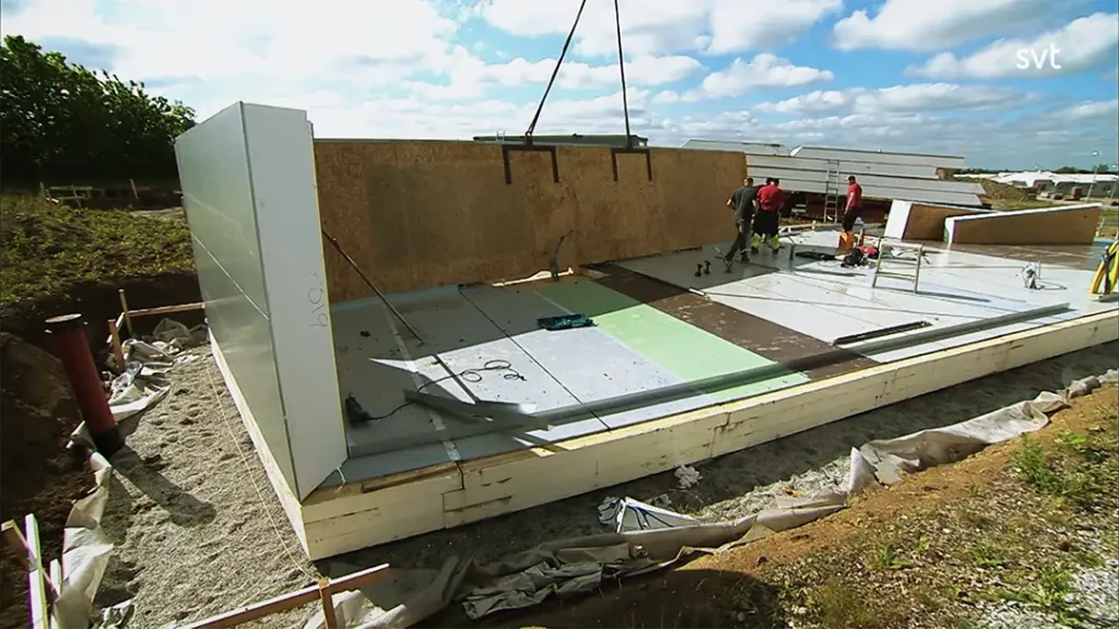 Stiligt - Byggnadsarbetare monterar prefabricerade väggpaneler på en husgrund på en byggarbetsplats under en delvis molnig himmel.