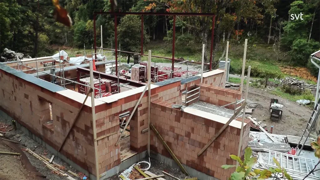 Stiligt - Delvis byggd byggnad med röda tegelväggar, byggnadsställningar och stålbjälkar, omgiven av byggmaterial och träd i bakgrunden.