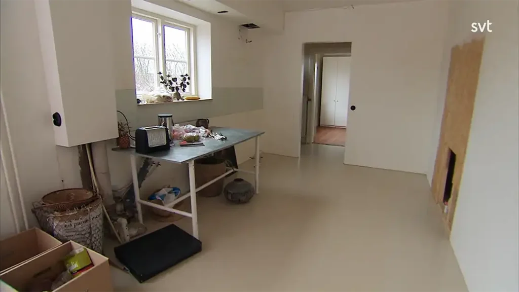 Stiligt - Ett sparsamt möblerat rum med ett bord med köksartiklar, ett fönster, en öppen dörröppning och lådor på golvet. Väggar och golv är ljusa.