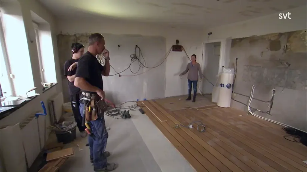 Stiligt - Tre personer står i ett delvis renoverat rum med ofullbordade väggar, synliga ledningar och delvis installerade trägolv. Byggverktyg och material är utspridda.