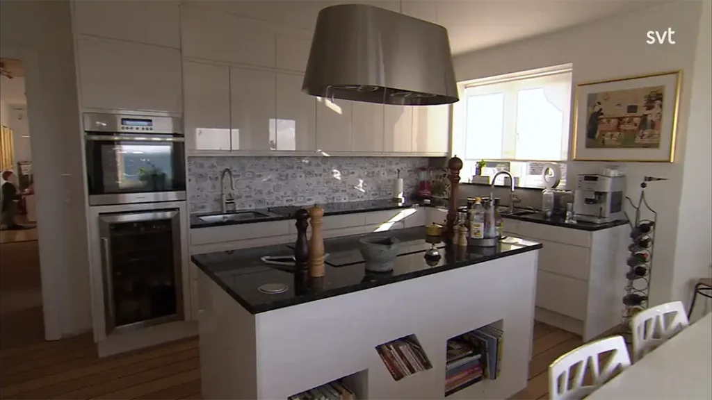 Stiligt - Modernt kök med vita skåp, en central ö med bokhyllor, vitvaror i rostfritt stål och en stor spiskåpa i metall. Solljuset strömmar in genom ett fönster ovanför diskbänken.