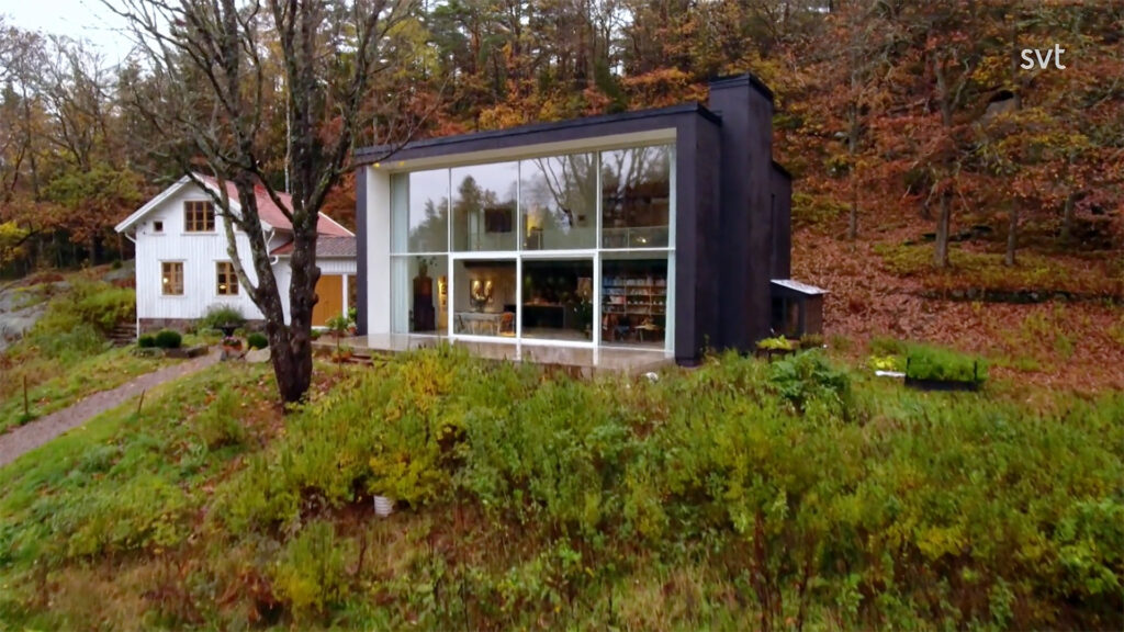 Stiligt - Ett modernt svart hus med stora fönster från golv till tak ligger bredvid ett traditionellt vitt hus, omgivet av höstträd och grönska.