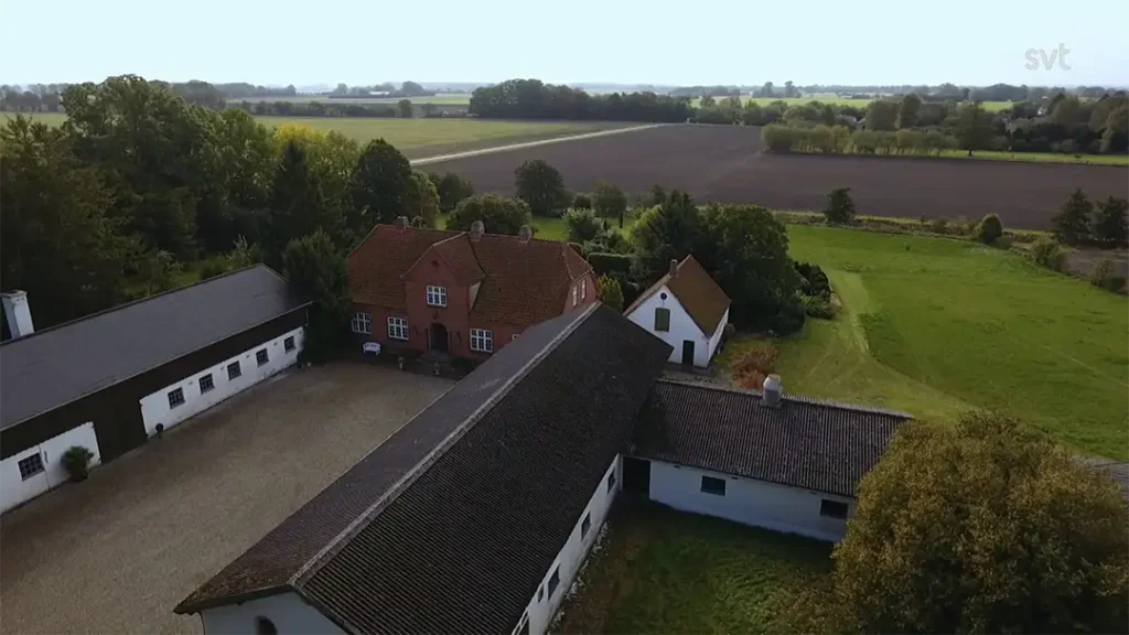 Stiligt - Flygfoto över en lantgård med rött tegel och vita byggnader omgivna av åkrar och träd under en klar himmel.