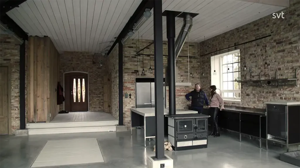 Stiligt - Två personer står och pratar i ett modernt kök i industriell stil med exponerade tegelväggar, stora fönster, metallbjälkar och minimalistisk inredning.