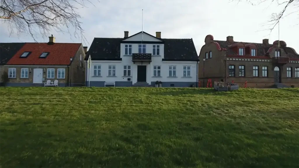 Stiligt - Tre hus står sida vid sida på en gräsbevuxen gräsmatta; det centrala huset är vitt med svarta detaljer, flankerat av tegelhus med röda tak.