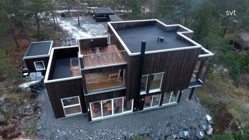 Stiligt - Flygfoto över ett modernt tvåvåningshus med platt tak, stora fönster och mörka träpaneler, beläget på en stenig tomt omgiven av träd.