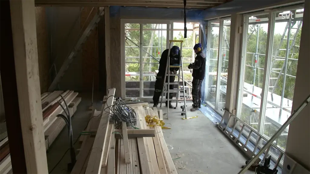 Stiligt - Två byggnadsarbetare i säkerhetsutrustning står nära ett fönster inne i en byggnad under uppbyggnad, med träbjälkar och verktyg utspridda på golvet.