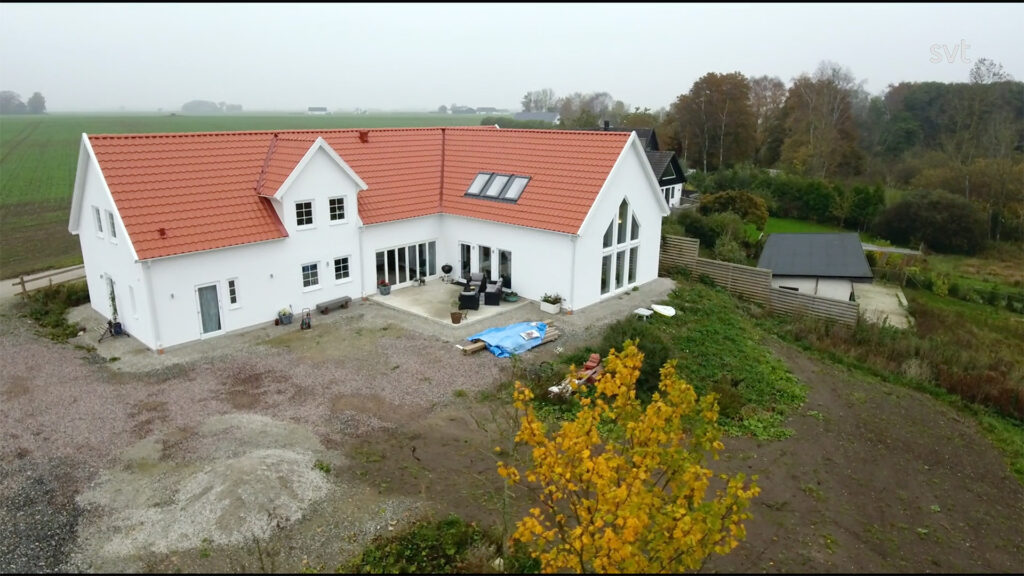 Stiligt - Ett stort vitt hus med rött tegeltak ligger på en rymlig tomt omgiven av åkrar och träd, med utemöbler och material synliga nära byggnaden.