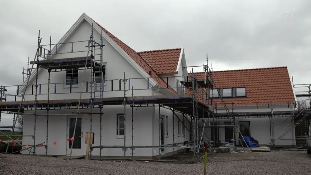Stiligt - Ett vitt hus med ett rött tegeltak under uppbyggnad, omgivet av byggnadsställningar en molnig dag.