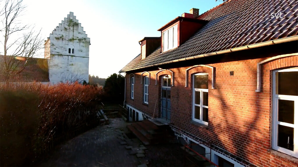 Stiligt - Ett tegelhus med välvda fönster står bredvid ett vitt gaveltorn under en klar himmel, med solljus som kastar skuggor på byggnaderna.