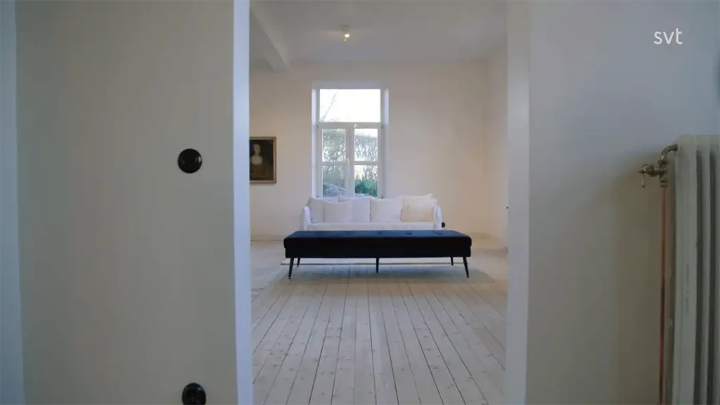 Stiligt - Ett minimalistiskt rum med en svartvit soffa, tre vita kuddar, en radiator, en inramad tavla på väggen och ett stort fönster som släpper in naturligt ljus.