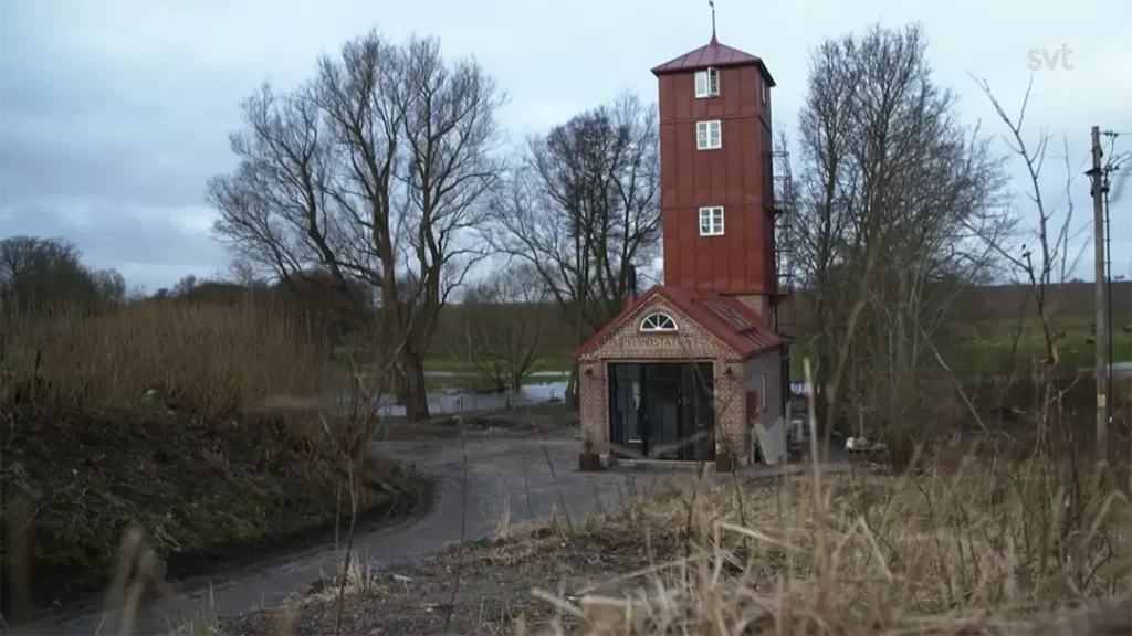Stiligt - En liten tornbyggnad i rött tegel och trä står bredvid en grusväg, omgiven av lövlösa träd och torrt gräs nära en flod under en molnig himmel.
