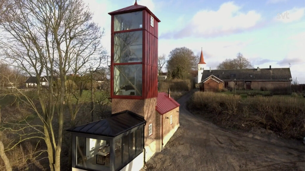Stiligt - Ett smalt tegelhus med ett högt glastorn står på en grusväg, omgivet av träd och lantliga byggnader, med en kyrka synlig i bakgrunden.