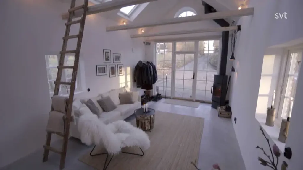 Stiligt - Ett modernt vardagsrum med vita väggar, en loftstege, en vit soffa, en matta, en vedspis och stora fönster som släpper in dagsljus.