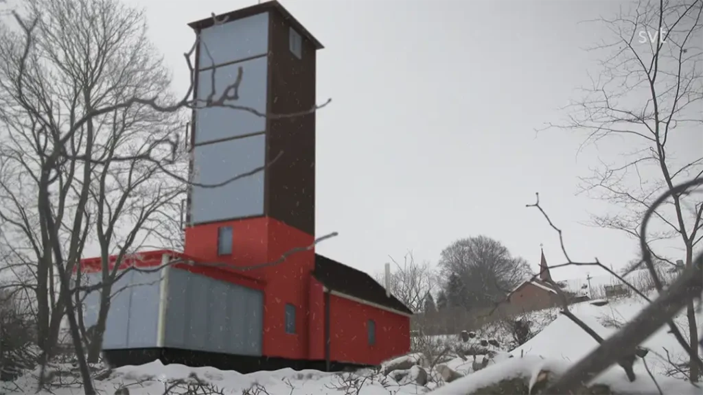 Stiligt - Ett högt, smalt hus med röda och blå ytterväggar står i ett snölandskap, omgivet av kala träd under en mulen himmel.
