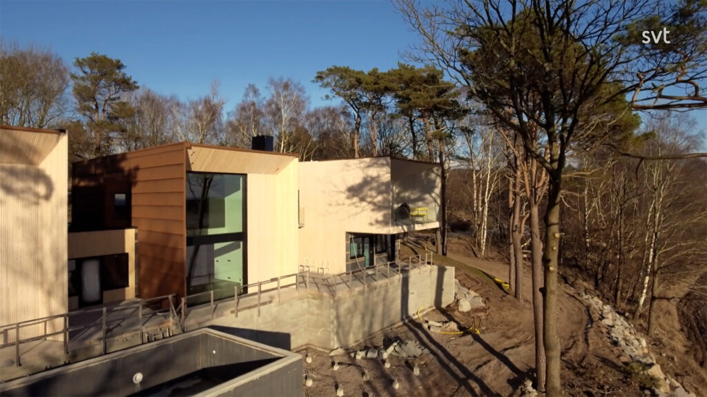 Stiligt - En modern, kantig byggnad med stora fönster ligger bland kala träd under en klar himmel. Konstruktionen verkar oavslutad med synlig betong och skräp.