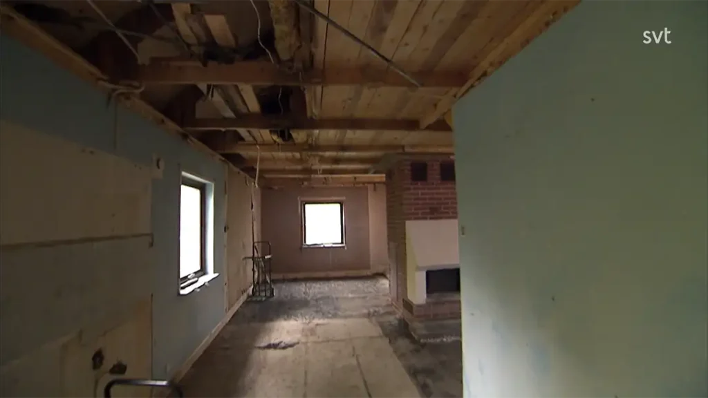 Stiligt - Ett rum under renovering med synliga takbjälkar i trä, oavslutade golv, ljusblå och vita väggar, två fönster och en öppen spis i tegel.