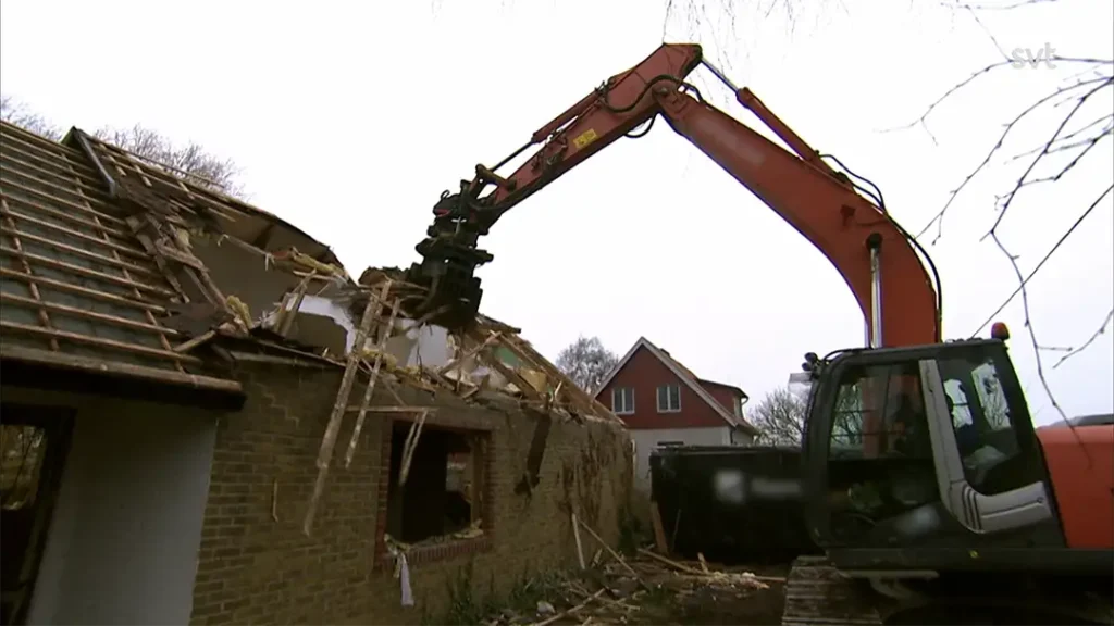 Stiligt - En grävmaskin river taket och väggarna på ett hus, med skräp som faller bredvid maskinen. Ett bostadshus syns i bakgrunden.