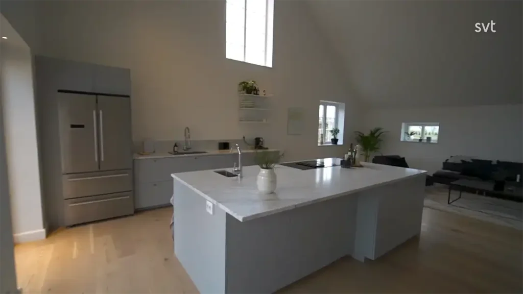 Stiligt - Modernt kök med en stor marmorö, kylskåp i rostfritt stål, ljusa trägolv och ett angränsande vardagsrum med soffa och växter. Dagsljuset kommer in genom höga fönster.