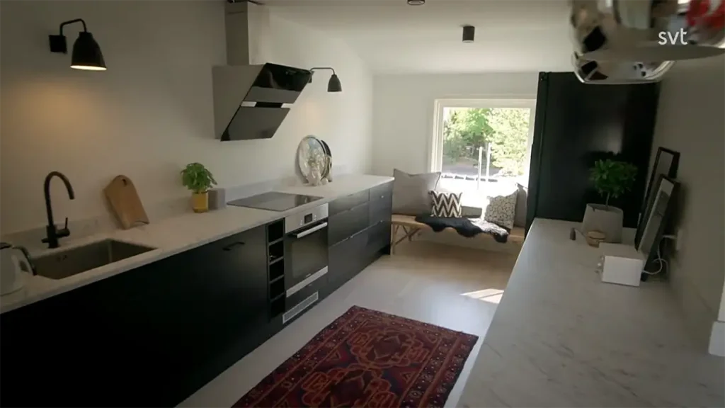 Stiligt - Modernt kök med svarta skåp, vita bänkskivor, en röd matta och en liten sittgrupp vid ett stort fönster med solljus som strömmar in.