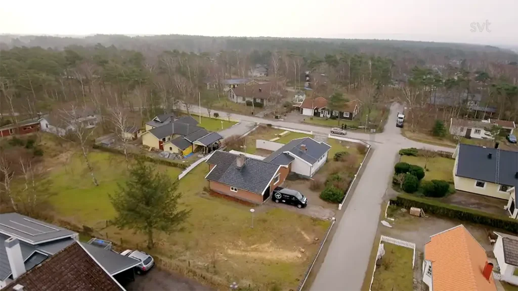 Stiligt - Flygfoto över ett bostadsområde med hus, några parkerade bilar och omgivande träd på en molnig dag.
