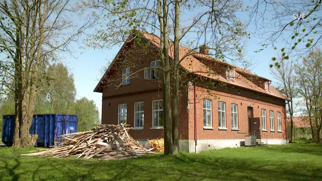 Stiligt - Ett tegelhus med rött tak och många fönster står på en gräsbevuxen gård, med en hög med träskräp och en stor blå soptunna i närheten.