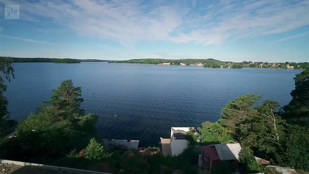 Stiligt - En bred sjö med lugnt blått vatten, omgiven av gröna träd och byggnader vid stranden, under en mestadels klar himmel med striga moln.