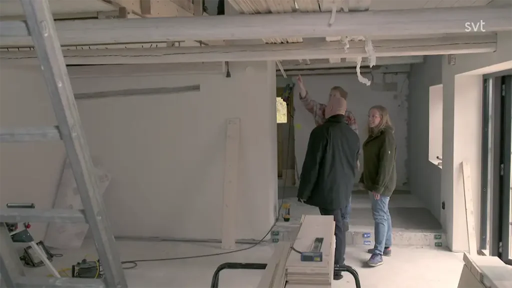 Stiligt - Tre personer står inne i ett delvis byggt eller renoverat rum och diskuterar interiören; byggmaterial och verktyg är synliga.