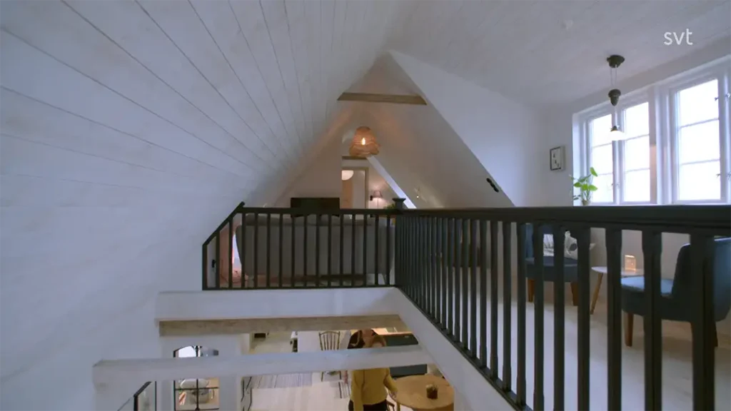 Stiligt - Ett modernt loft med snedtak i vit panel, svarta räcken, stora fönster, och utsikt över nedre våningen med en person som sitter vid ett bord.