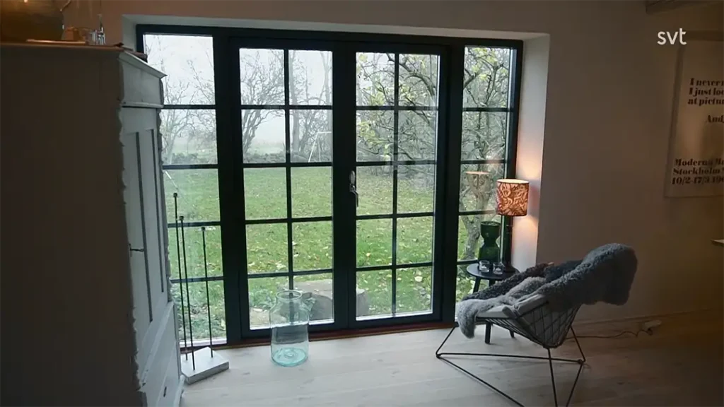 Stiligt - Ett modernt rum med ljusa trägolv och en glasdörr med svart inramning har utsikt över en gräsbevuxen gård med träd. En stol, lampa och ett litet bord är placerade vid dörren.