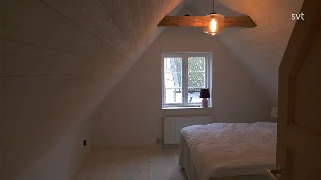 Stiligt - Ett litet vindsrum med sneda trätak, en dubbelsäng, ett fönster med en lampa på tröskeln och en taklampa.