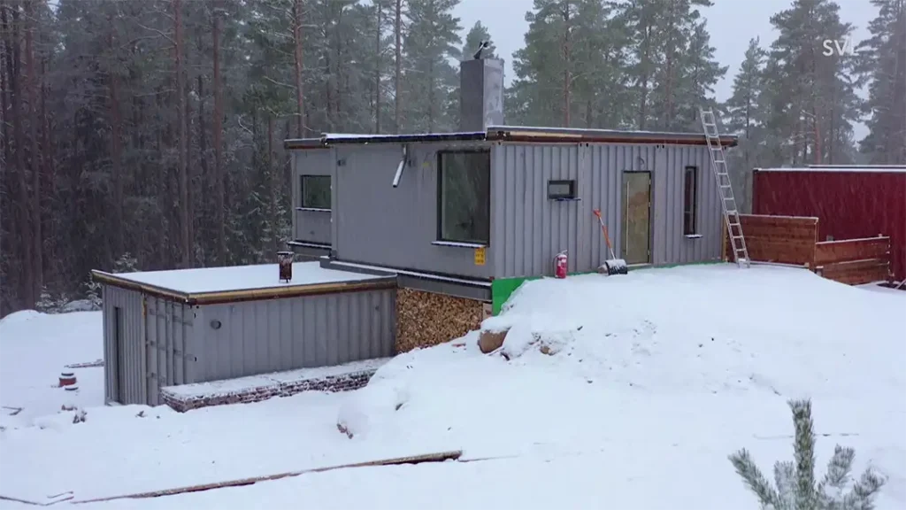 Stiligt - Ett modernt, rektangulärt, grått hus med platta tak står i ett snölandskap omgivet av tallar; en stege lutar mot sidan.