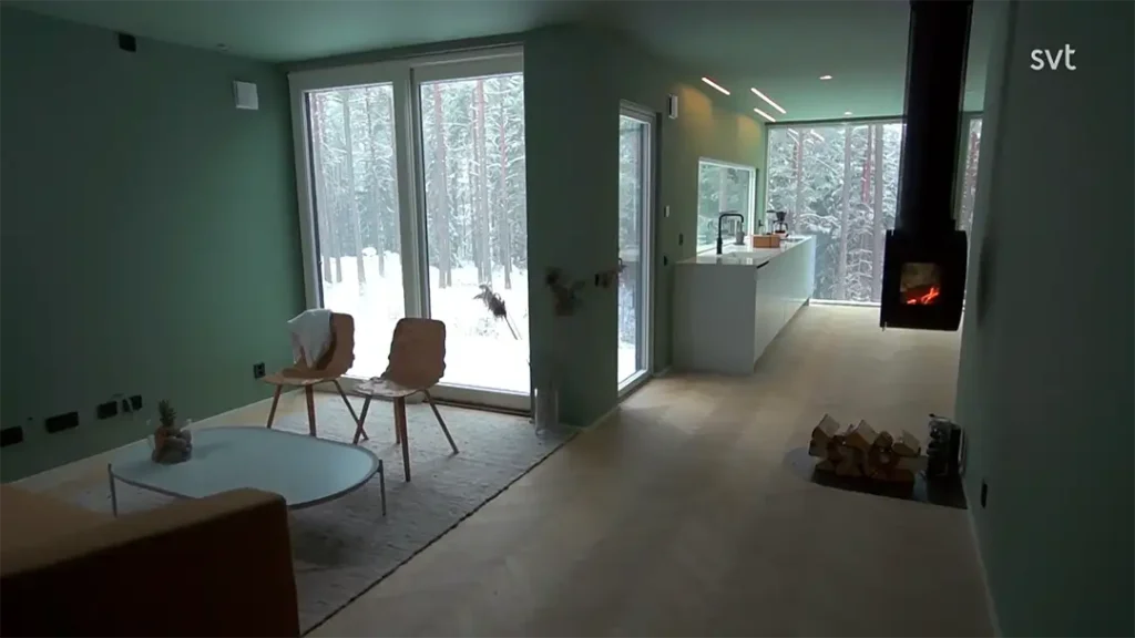 Stiligt - Modernt vardagsrum och kök med stora fönster som visar en snöig skog, ljusgröna väggar, trästolar, ett soffbord och en tänd upphängd öppen spis.