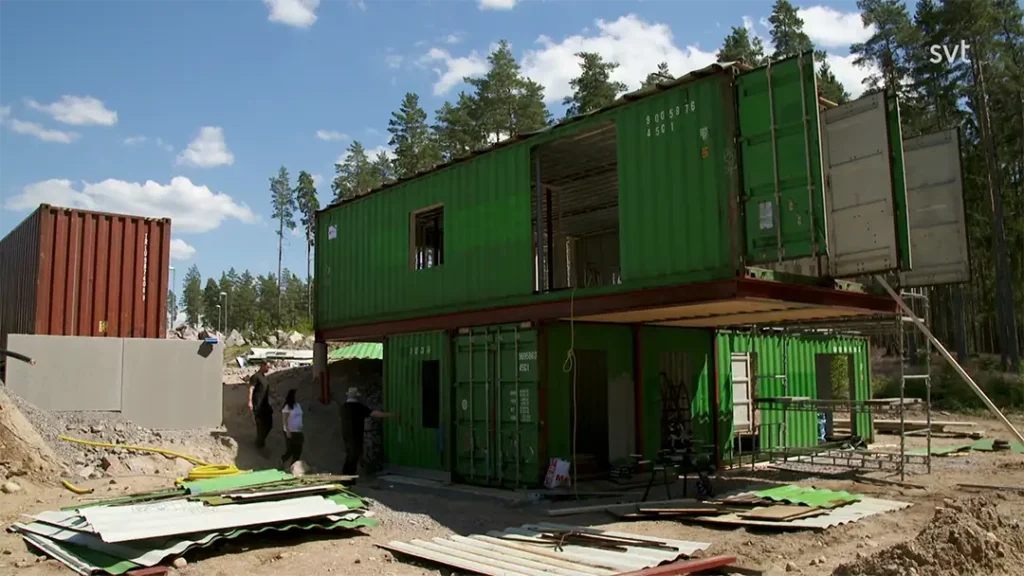 Stiligt - Två gröna fraktcontainrar staplas och görs om till en byggnad under uppbyggnad på en trädbevuxen plats, med byggmaterial utspridda.