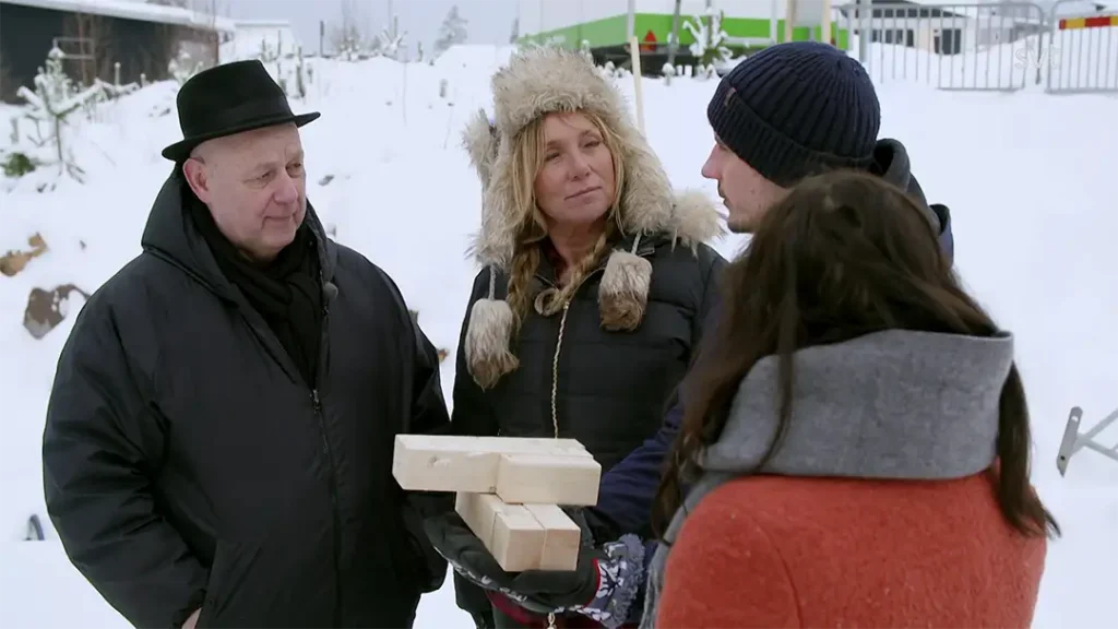 Stiligt - Fyra personer står ute i snöväder, två håller i träbitar, alla varmt klädda i vinterrockar och hattar.