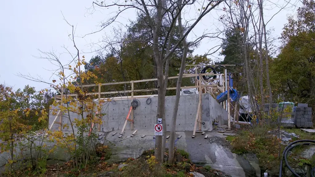 Stiligt - En betongkonstruktion under uppbyggnad sitter på en stenig yta, omgiven av träd. Träräcken och byggmaterial är synliga, med en "ingentréskylt" på staketet.