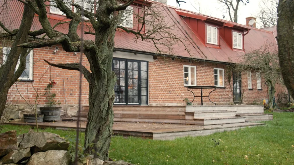 Stiligt - Ett tegelhus med rött tak har ett trädäck, svarta glasdörrar och ett runt bord med ett bladlöst träd i förgrunden.