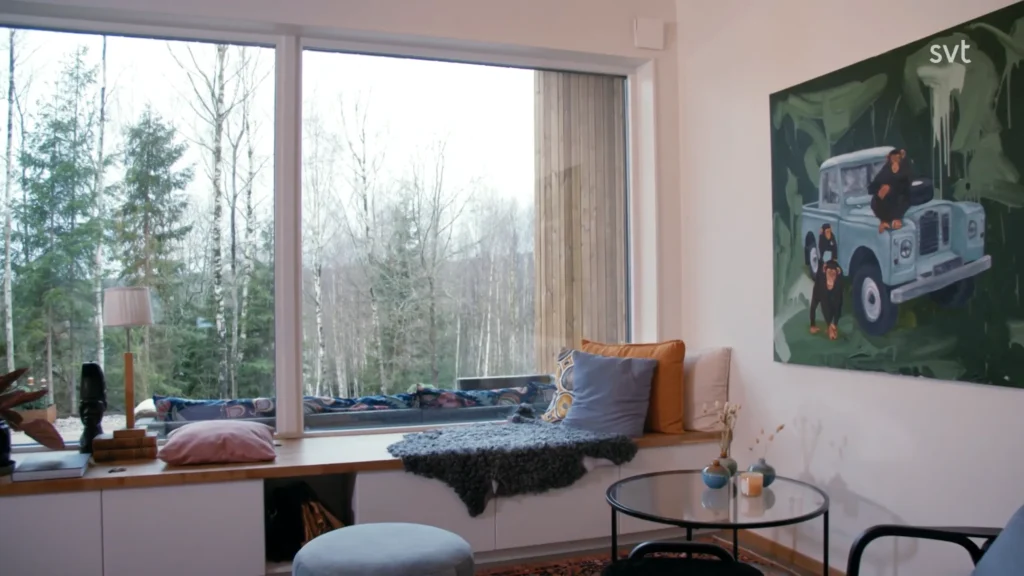 Stiligt - Ett mysigt rum med ett stort fönster med utsikt över träd, en bänk med kuddar, ett litet runt bord och en målning av en bil på väggen.