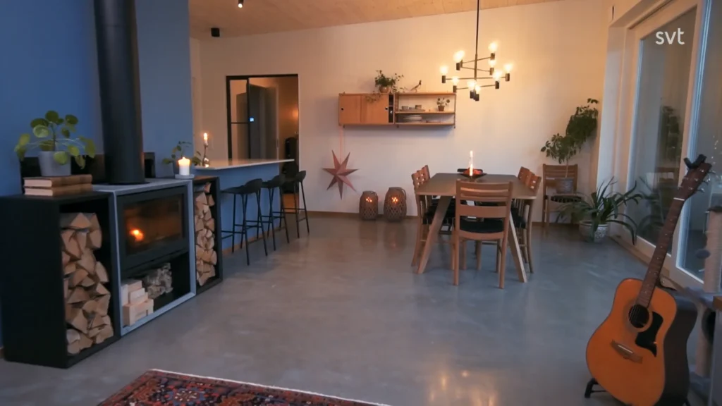 Stiligt - Modern mat- och vardagsrumsdel med vedspis, staplad ved, matbord, hyllor, krukväxter, gitarr och varm belysning.