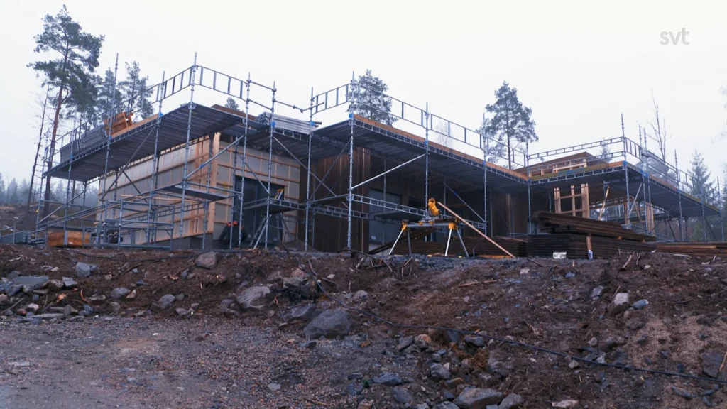 Stiligt - Ett modernt hus under uppförande är omgivet av byggnadsställningar på en stenig, lerig plats med några träd i bakgrunden.