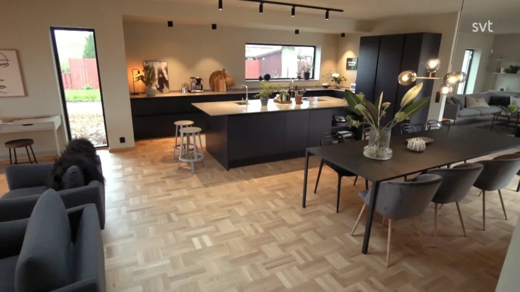 Stiligt - Modernt kök och matplats i öppen planlösning med svarta skåp, stor ö, parkettgolv och minimalistisk inredning; vardagsrum synlig i bakgrunden.