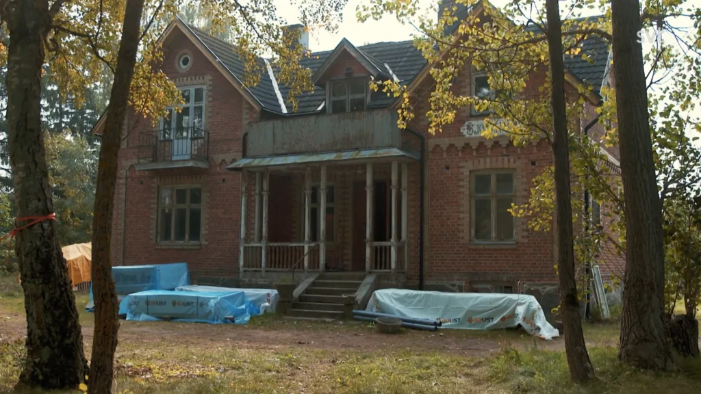Stiligt - Ett tvåvåningshus i tegel under renovering, med byggmaterial och utrustning täckt av blå presenningar på gården omgiven av träd.