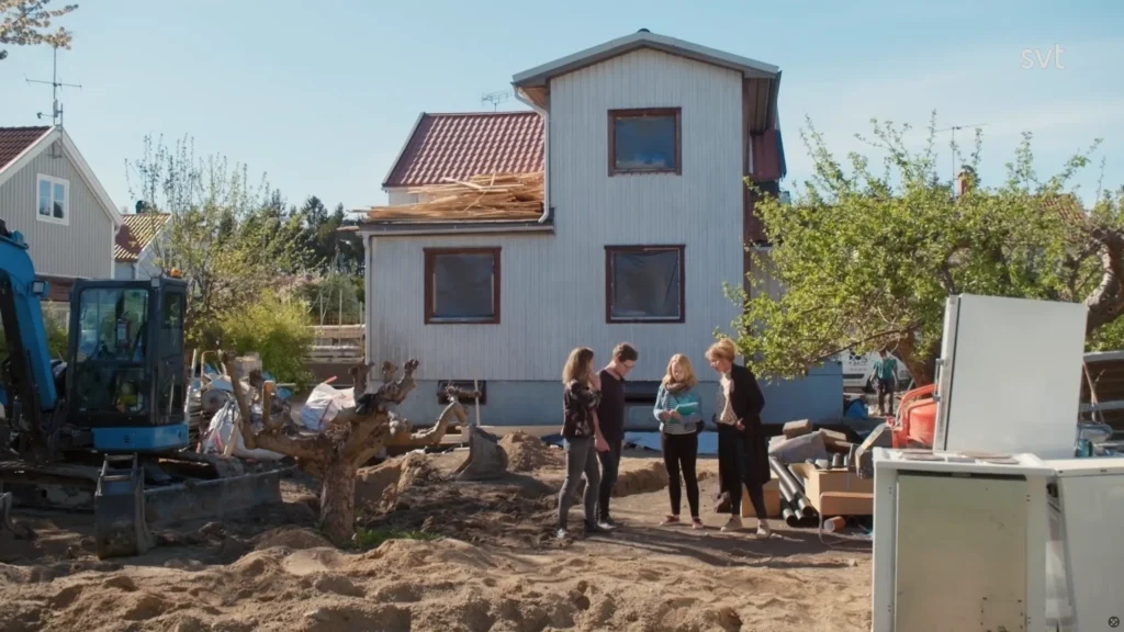 Stiligt - Fyra personer står och pratar på en gård fylld med byggmaterial och smuts framför ett delvis renoverat tvåvåningshus med skadat tak.