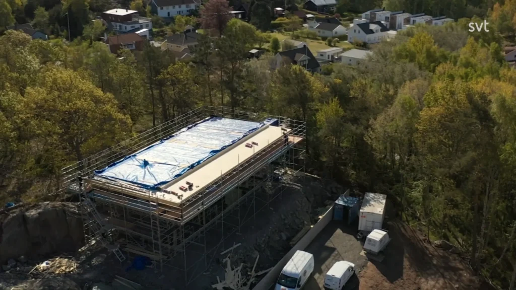 Stiligt - Flygfoto över en rektangulär byggnad under uppbyggnad, täckt med en presenning, omgiven av byggnadsställningar och träd, med flera fordon parkerade i närheten i ett bostadsområde.