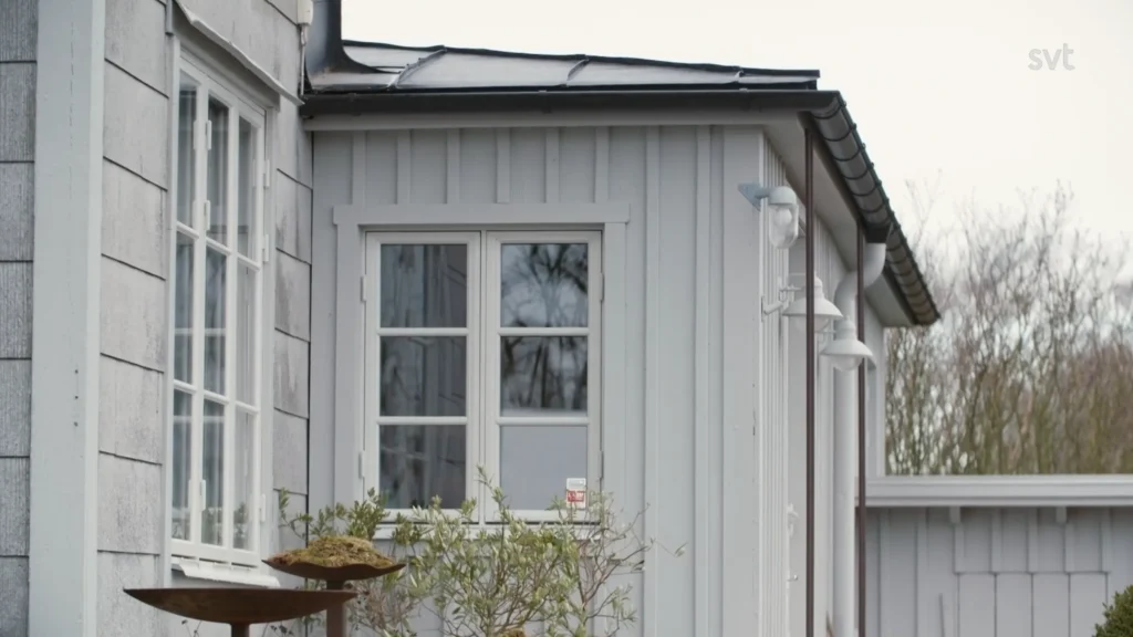Stiligt - Ljusgrått trähus exteriör med vittrimmade fönster, en liten växt och en fågelmatare i förgrunden; "svt" logotyp i det övre högra hörnet.