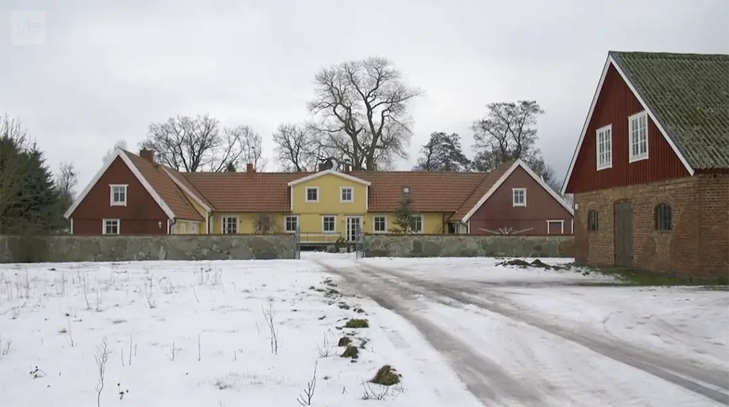 Stiligt - En stor gul bondgård med röda sidobyggnader och en ladugård i tegel, omgiven av en snöig gård och kala träd under en molnig himmel.