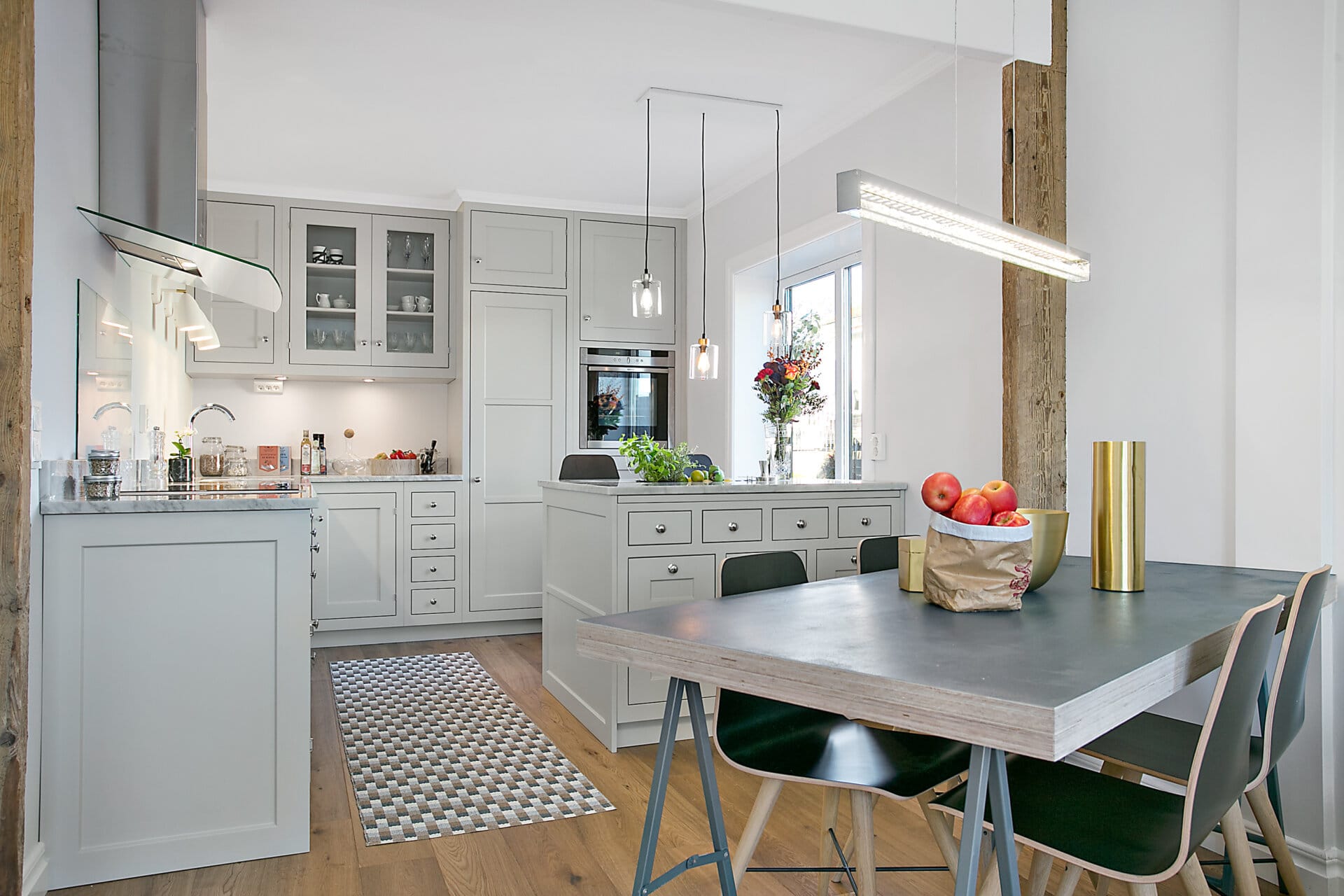 Stiligt - Modernt kök och matplats med ljusgrå skåp, svart matbord, fyra stolar, trägolv, pendelbelysning, samt ett fönster med blommor på tröskeln.
