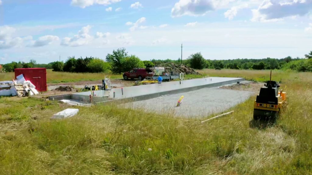 Stiligt - En betonggrundsplatta byggs på en gräsbevuxen tomt, med byggmaterial, ett rött fordon och utrustning synlig i närheten under en delvis molnig himmel.